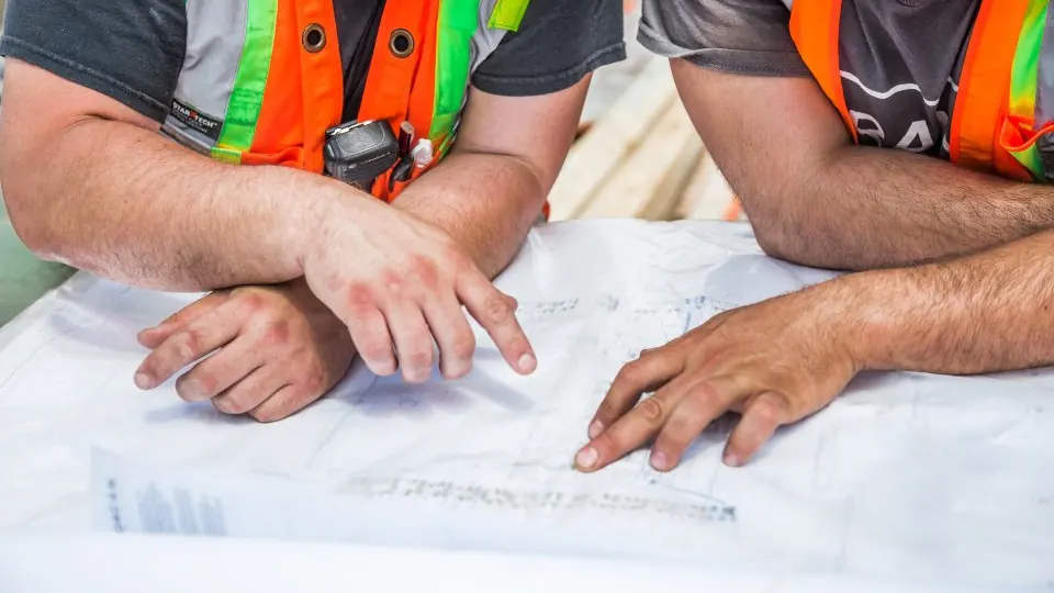 Two people in high-visibility vests reading a set of building drawings spread on a table