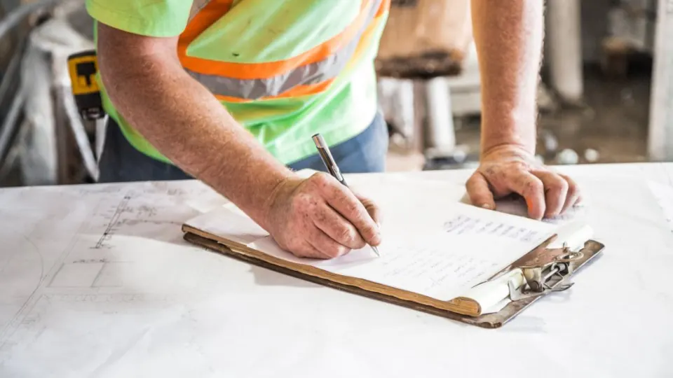 A hand writing a line-by-line schedule of work on a clipboard resting on rolled drawings