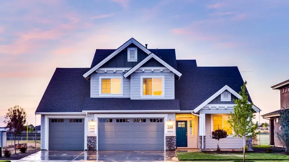 A two-storey suburban family home at dusk with lit windows, a pitched asphalt-shingle roof and an attached double garage