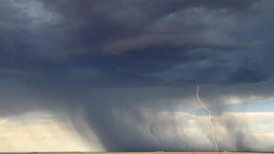 A storm cell trailing a shaft of rain across flat open country, with a lightning strike at the horizon
