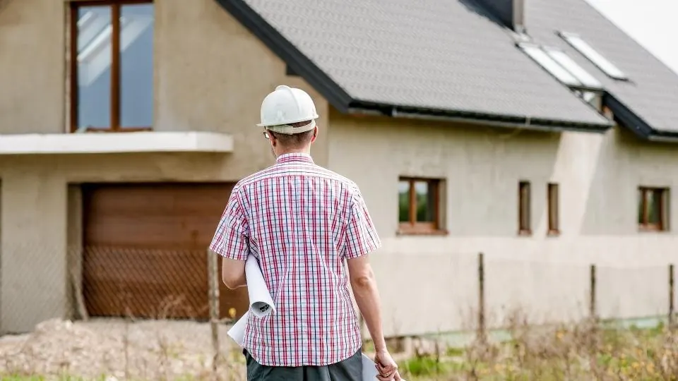 A person in a hard hat carrying rolled drawings, walking towards a house to survey it