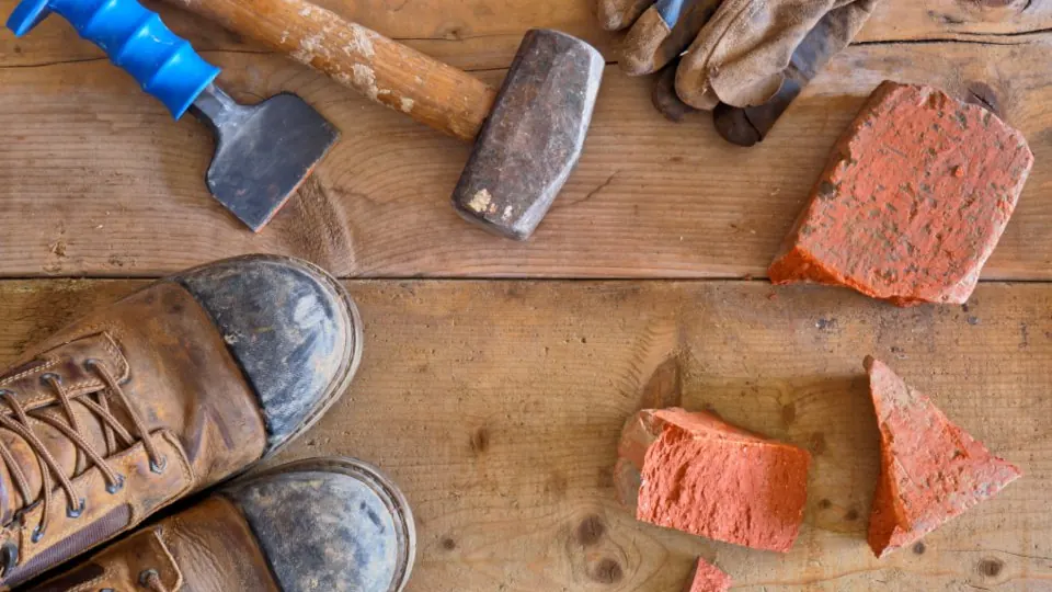 Hammers, work gloves, boots and broken brick arranged on a timber surface